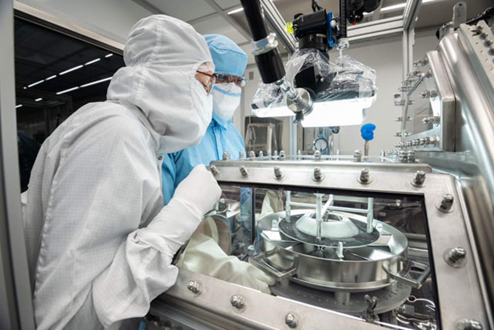 Two scientists wearing protective suits and face masks examine an asteroid sample inside a glovebox.